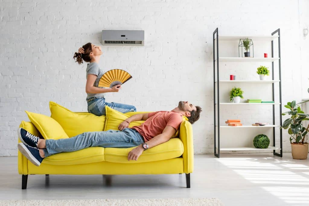 Licensed, Bonded, and Insured HVAC Specialists in King & Pierce Counties | exhausted man lying on yellow sofa under air conditioner near woman with hand fan